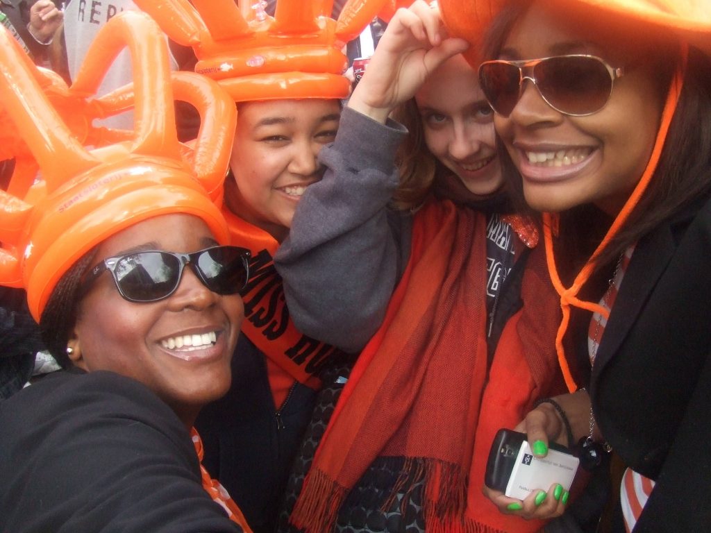 Four people smile at the camera. All are wearing orange headpieces made of balloons.