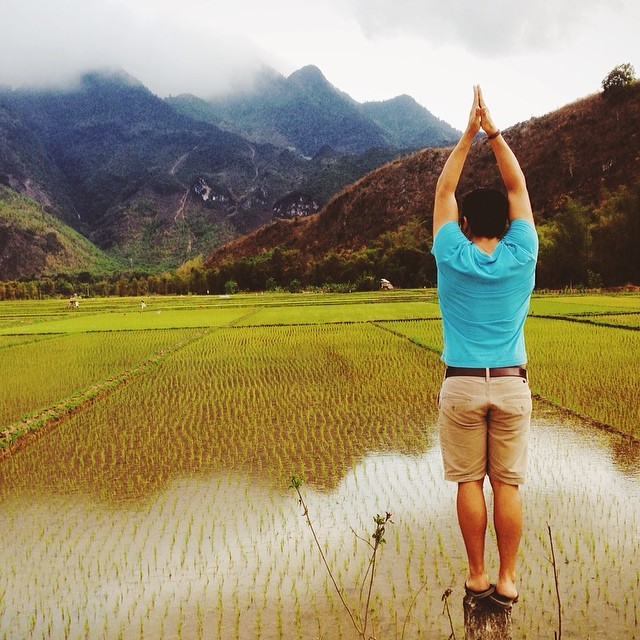 A student, with back to the camera, strikes a yoga warrior pose while standing in a green field of rice paddies in Vietnam. Mountains are in the background.