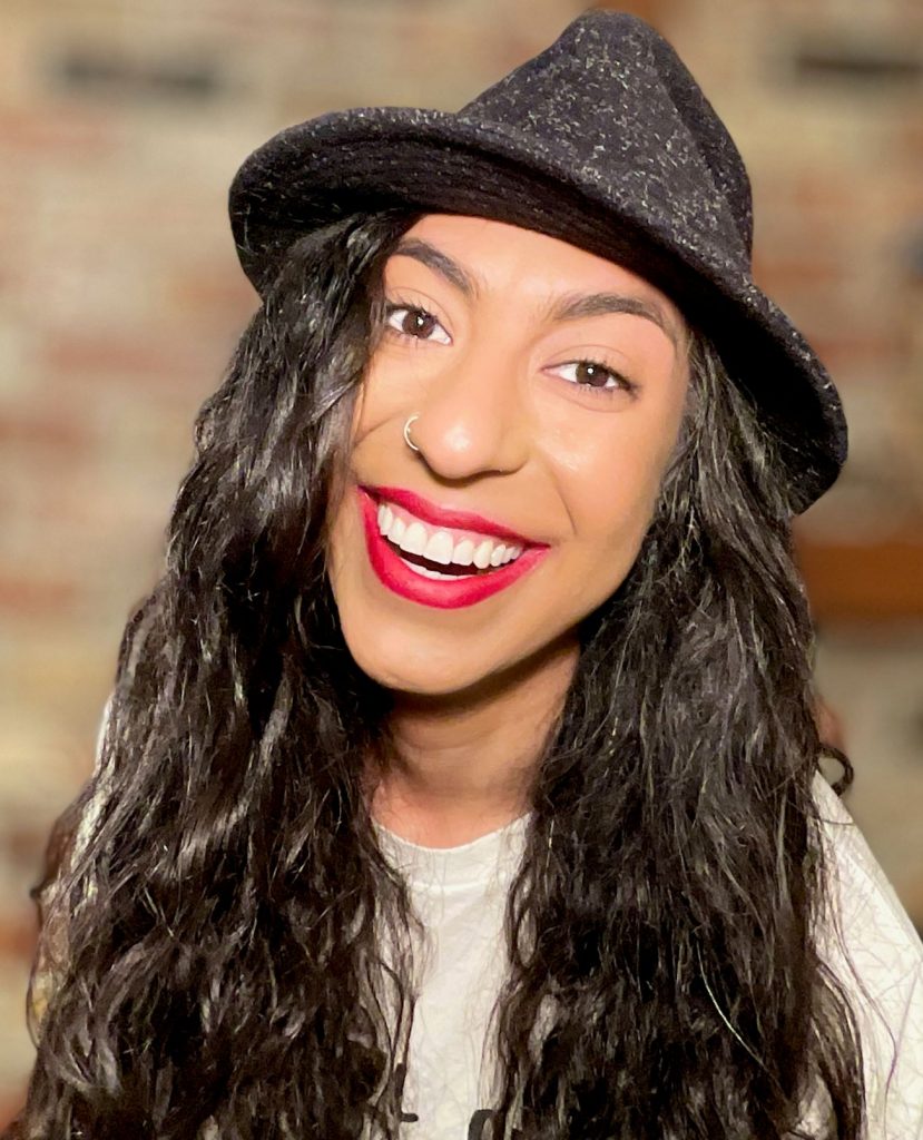 A woman with long dark hair, a small nose ring, bright lipstick and a brown fedora-style hat smiles broadly.