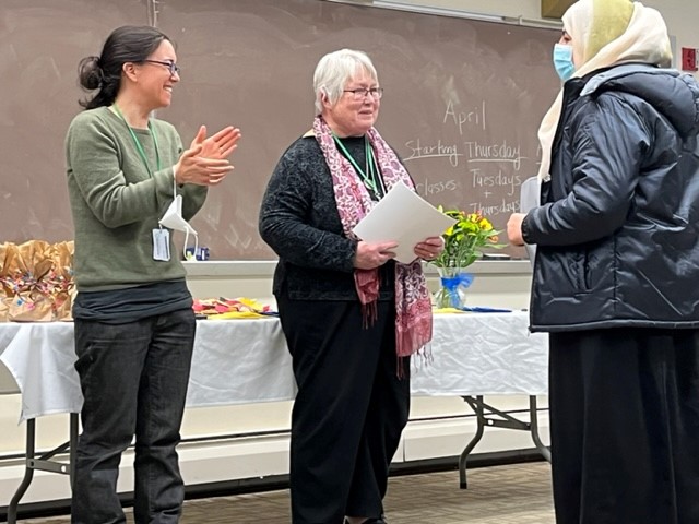 Two teachers, one clapping and the other holding papers, smile at a woman wearing a light-colored headscarf, a surgical mask, and a dark parka.