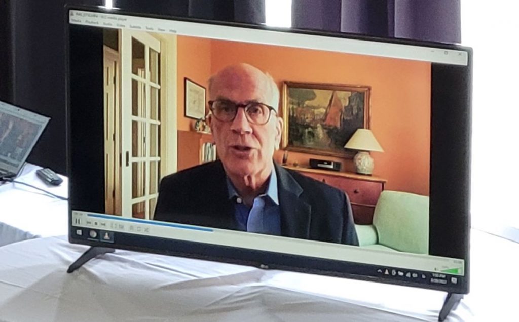 A man facing forward and speaking, featured on a TV screen