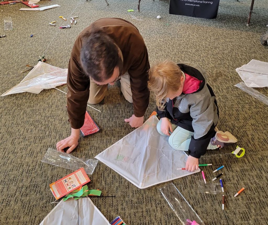 A man and child on the floor making a kite. The child holds a colored marker.