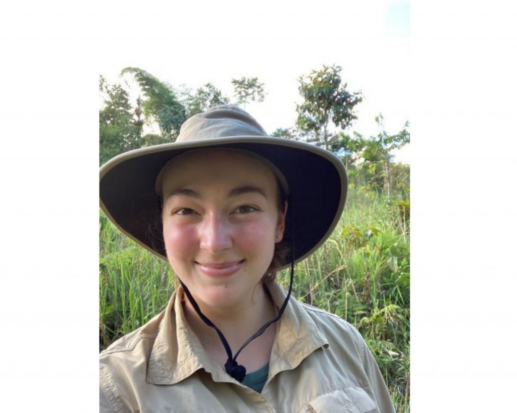 A young woman wearing a broad-brimmed safari hat and khaki-colored shirt stands against a tropical backdrop.