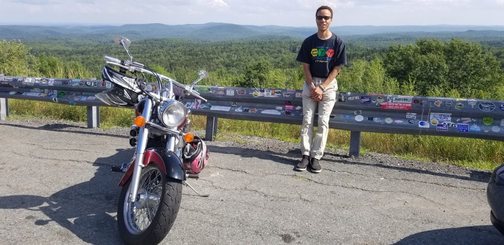 A man leans against a guardrail. A motorcycle is parked nearby. The background is green and mountainous.