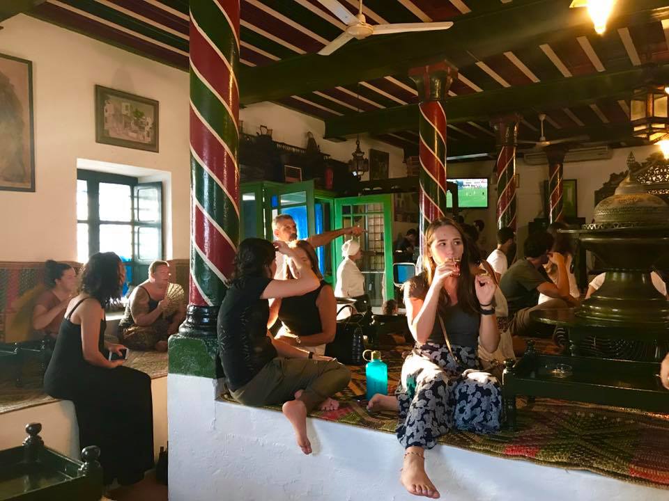 Men and women sitting in a Tunisian shop with white walls, a red and green poll and a white ceiling fan