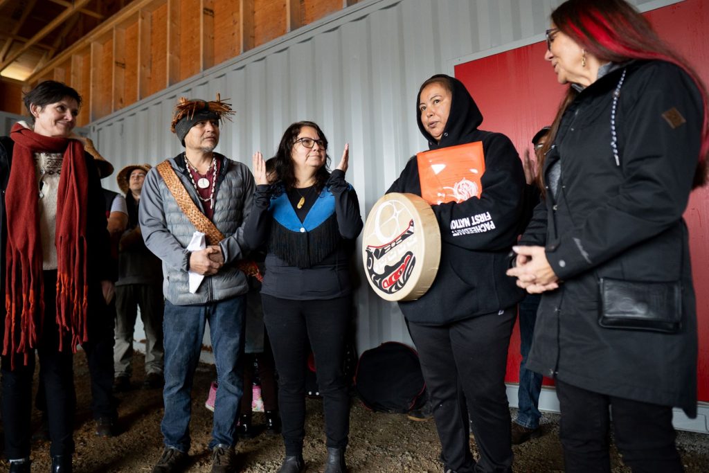 Five people are shown, including one holding an Indigenous drum