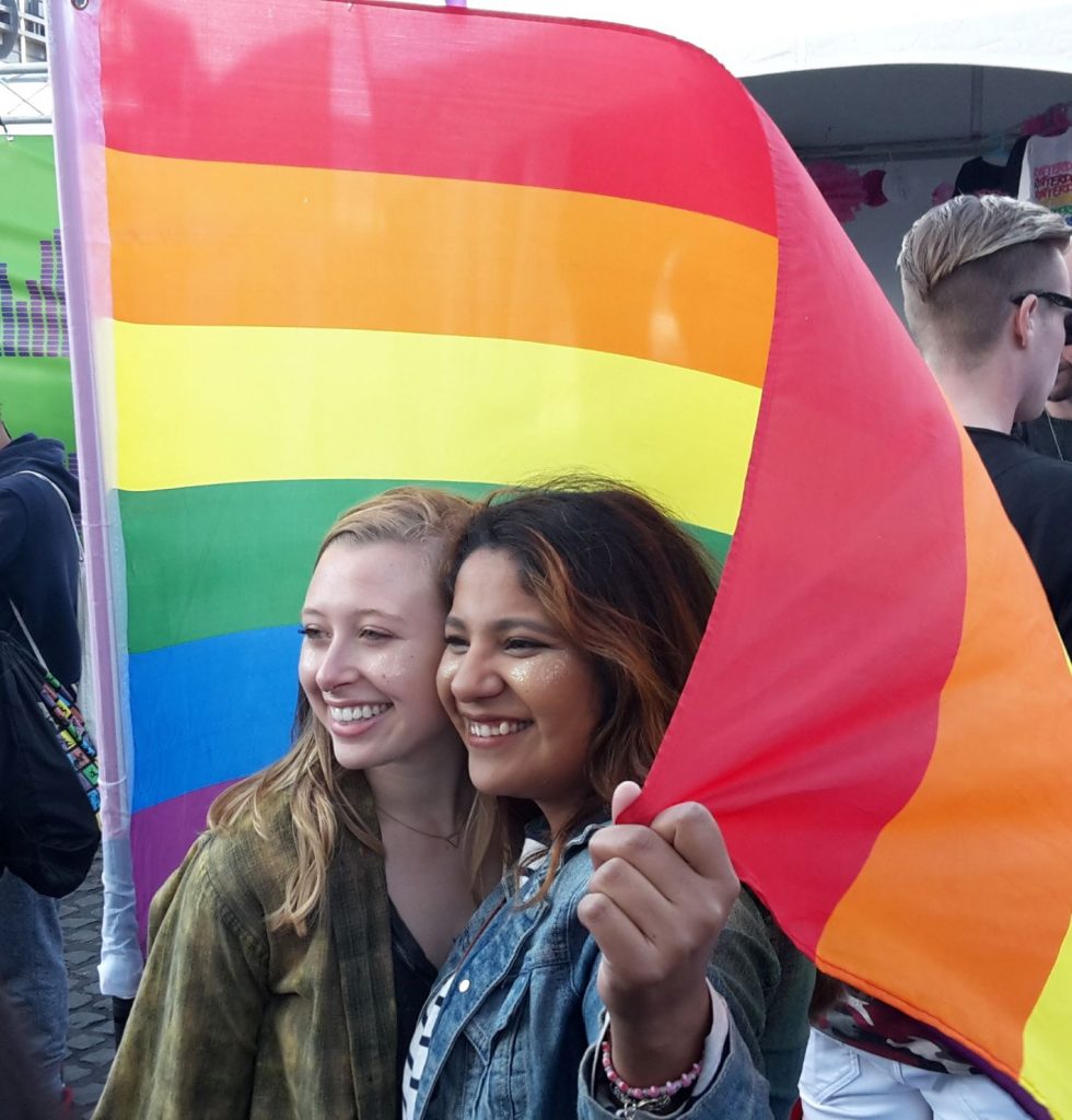 Two young woman smile as they are encircled by a rainbow flag.