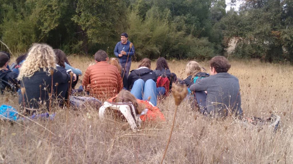 A group of students sits in a field of dry grass, facing a man giving a lecture.