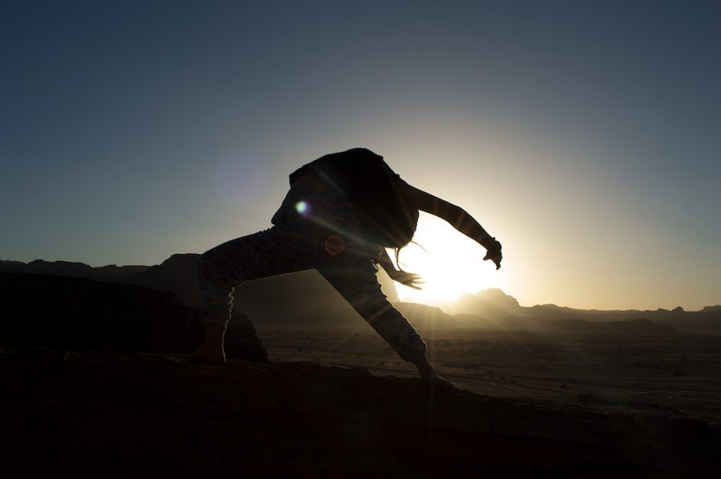 A student stretching backward is silhouetted against the setting sun in a desert landscape.