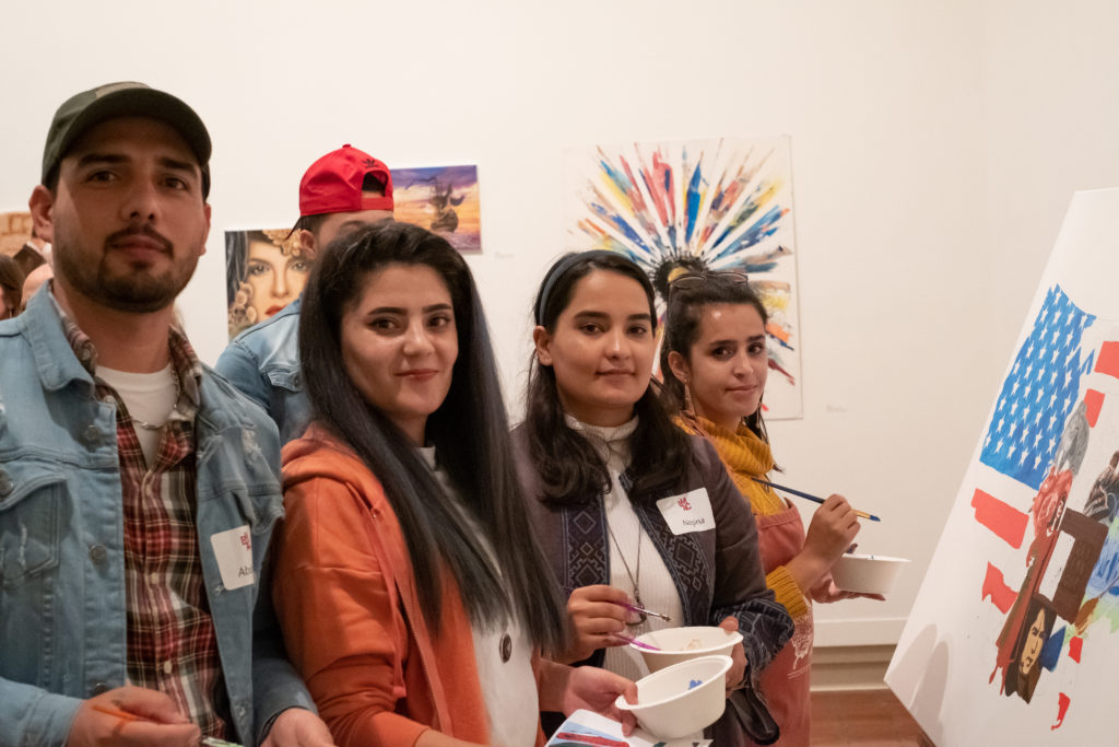 Four young people holding paint and paintbrushes, pose in front of a painting of a U.S. map with Afghan images.