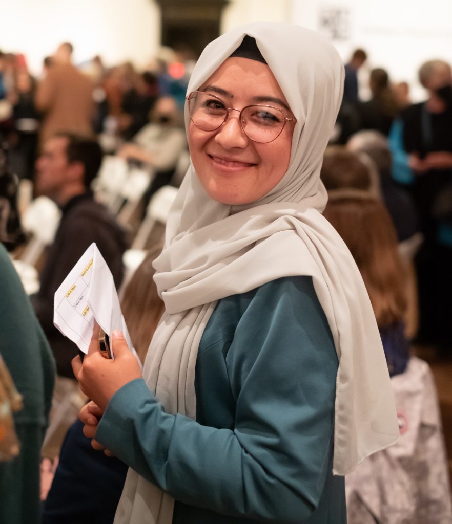 A woman smiles at the camera. She wears glasses, a long beige headscarf and a blue dress. She is holding a paper.