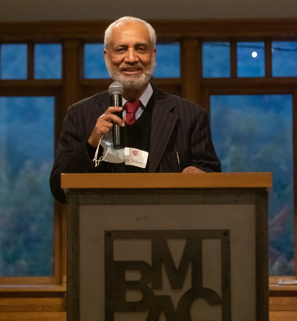 A man smiles as he speaks into a microphone at a podium. He has gray hair and a gray beard and wears a suit.