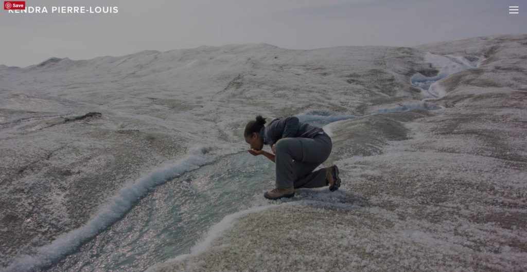 A woman kneels to drink drink from a stream of water running down a barren landscape.