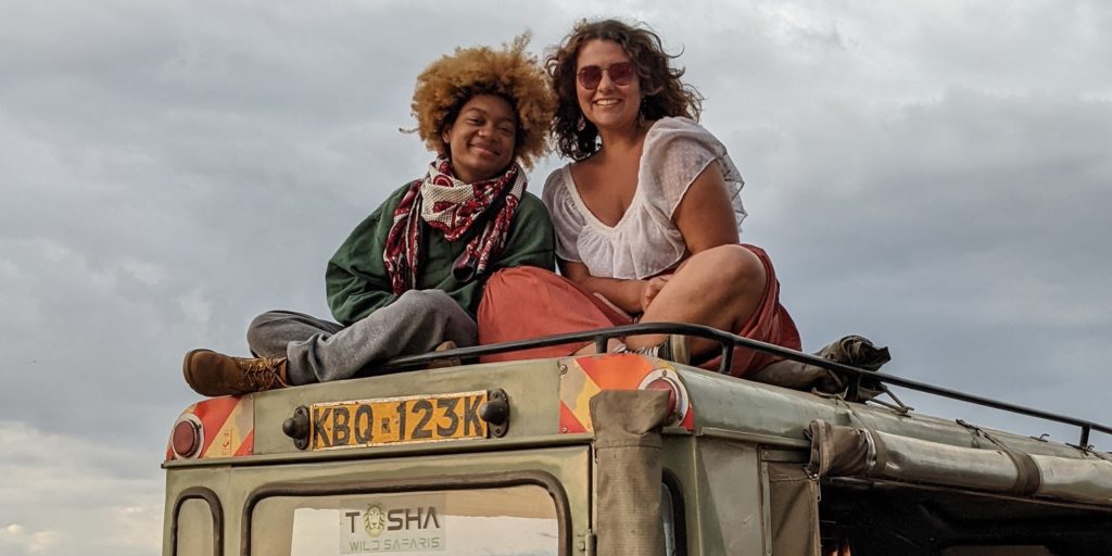 Two people atop a safari vehicle. One wears a scarf around their neck, a green sweatshirt, and gray pants. The other wears a white blouse. Both smile toward the camera.