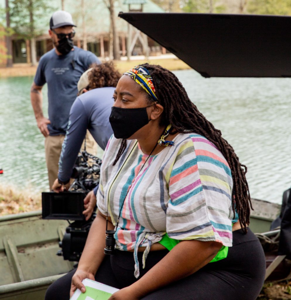 A woman with long dark hair, wearing a black surgical mask, on a film set. In the background is a camerman and another man. The set is on a body of water.