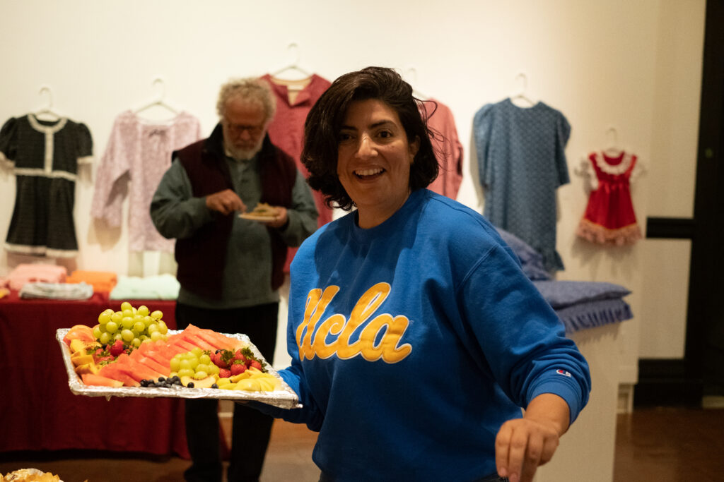 A woman with short dark hair, wearing a blue shirt, carries a tray of meats, cheese, and fruit.