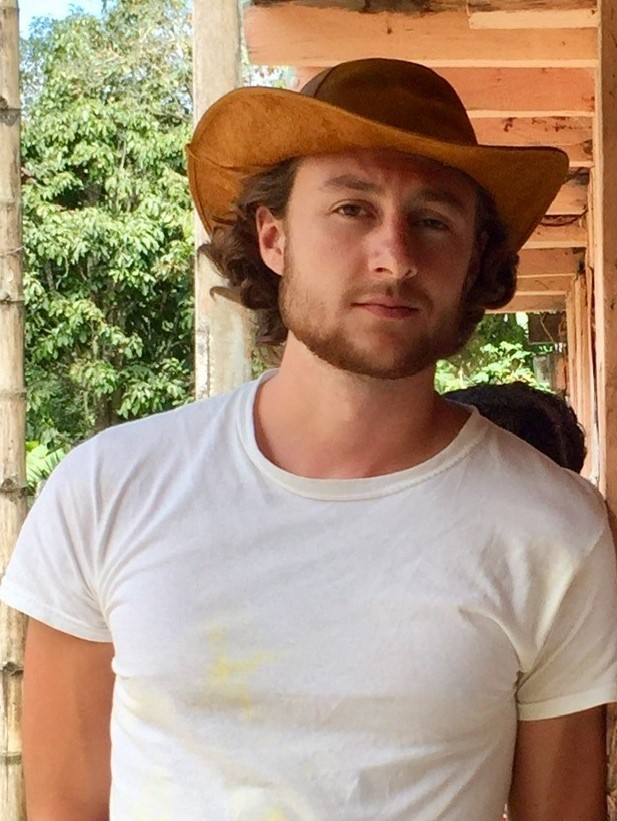 A man looks at the camera. He has light brown hair and a short beard, and is wearing a cowboy-type hat and a white T-shirt.