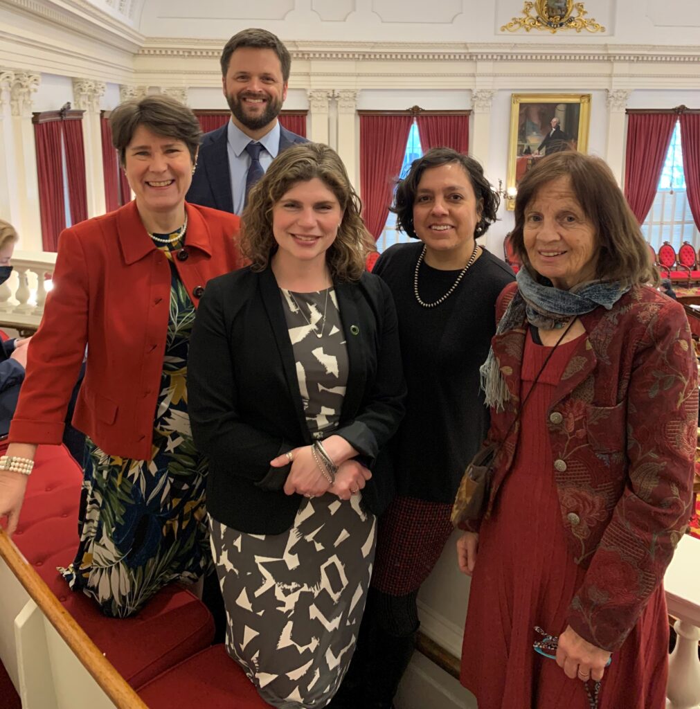 A group of four women and one man smile toward the camera. They are standing in an ornate room with red drapes and a gold-framed painting. 