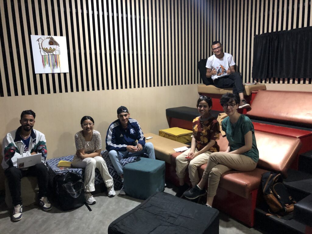 Six young people are seated on blue and red cushions and couches around a room. The wall has black stripes and a large TV screen.