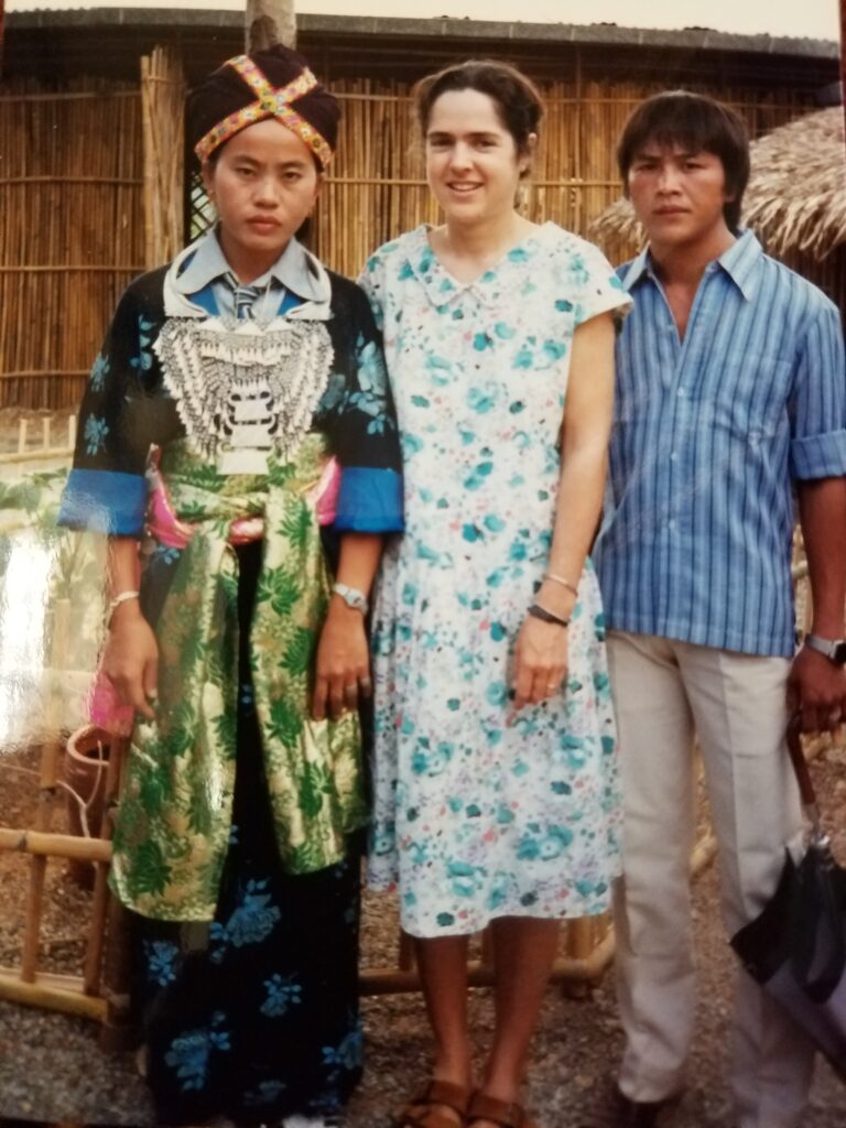 A young woman in a floral print dress stands between two people, one in ethnic dress and one in western-style clothing; blue button-down shirt and khaki pants.