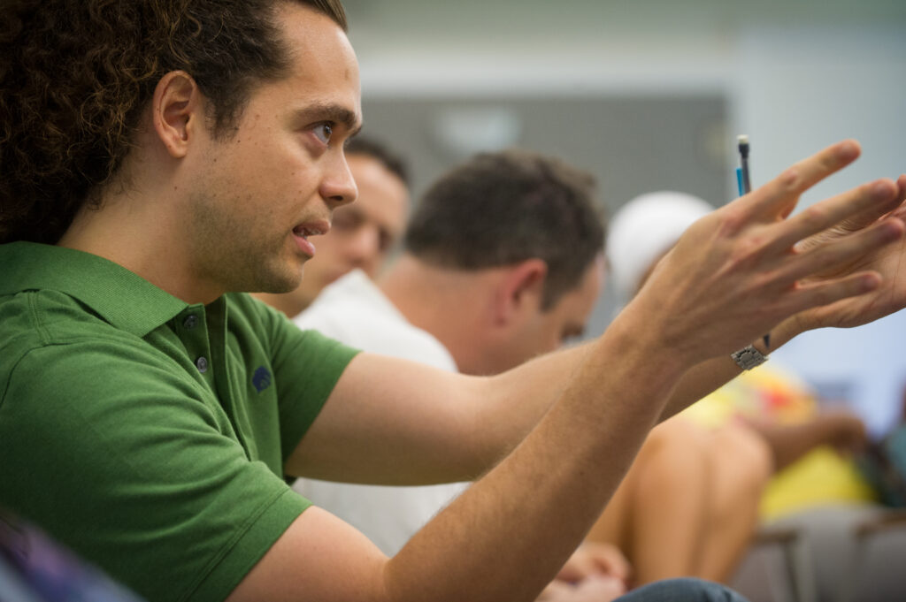 A young person with black hair and wearing a green polo shirt appears to be explaining something. His arms and hands are stretched forward.