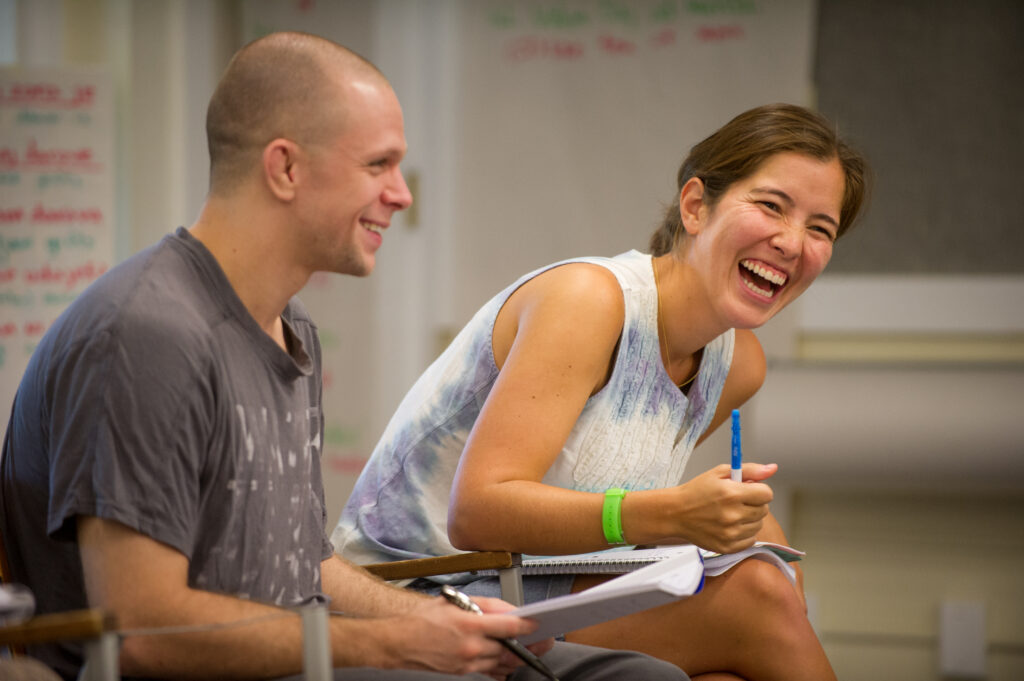 Two people seated in a classroom. The woman on the right leans forward laughing while the man on the left smiles.