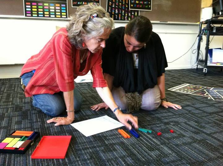 A woman wearing a red tunic and jeans, with salt-and-pepper shoulder-length hair, kneels on a carpeted floor next to a person in a black shirt. Both are looking at a white sheet of paper.