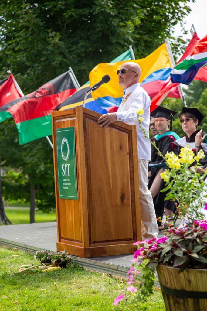 A man with shaved head and light gray beard, wearing sunglasses and a white embroidered tunic, stands at a podium with a sign reading "SIT"