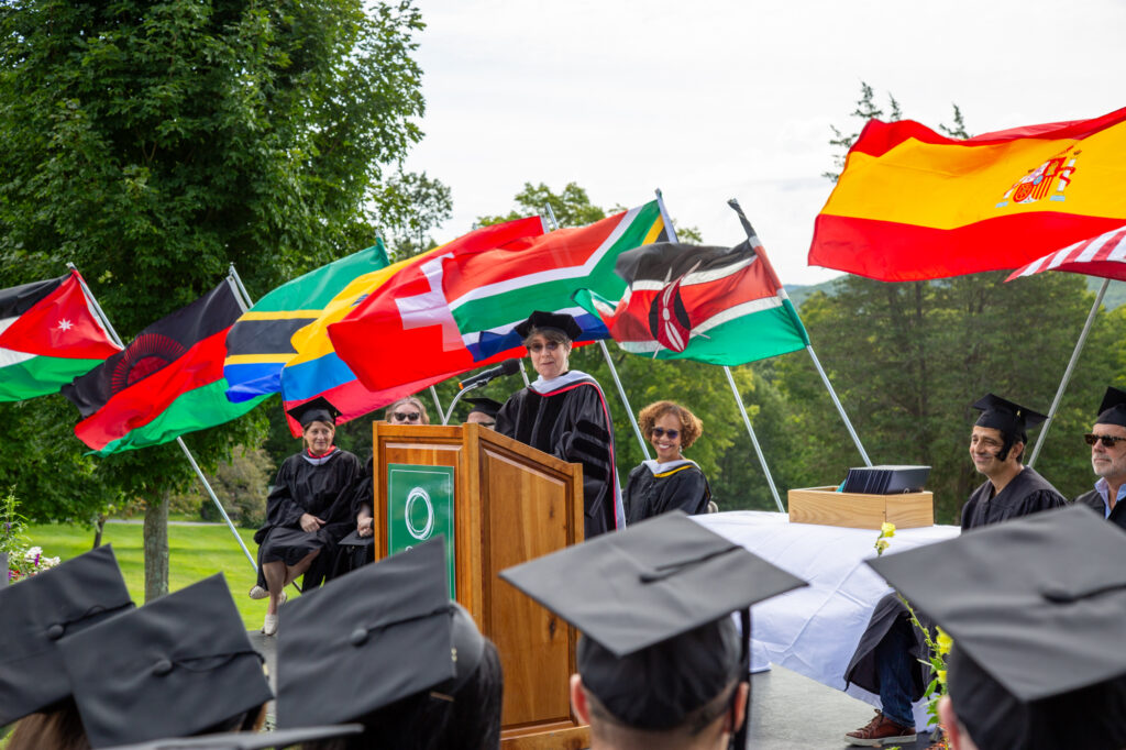 A women with short brown hair and sunglasses, wearing a PhD cap and black gown, stands at a podium facing a group of graduates whose caps can be seen in the photo. Behind her are various flags.