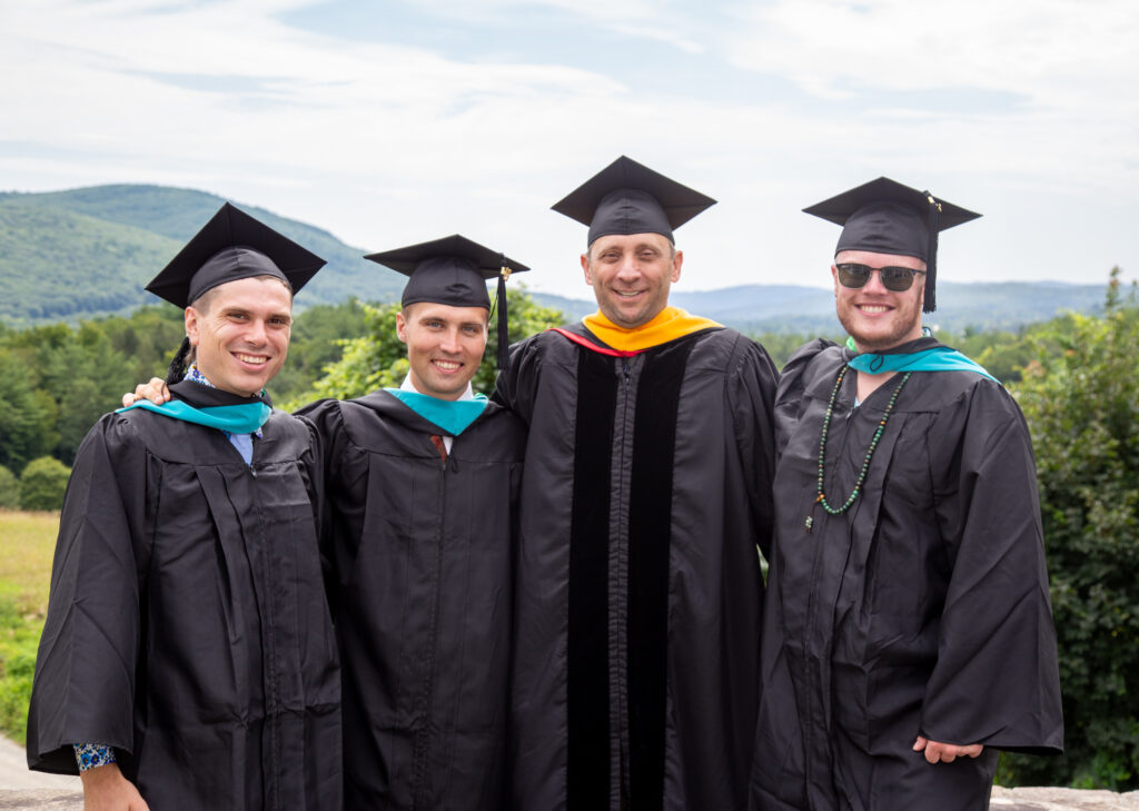 A group of four white men wearing black gowns and caps smile as they face the camera. In the background are green mountains and trees