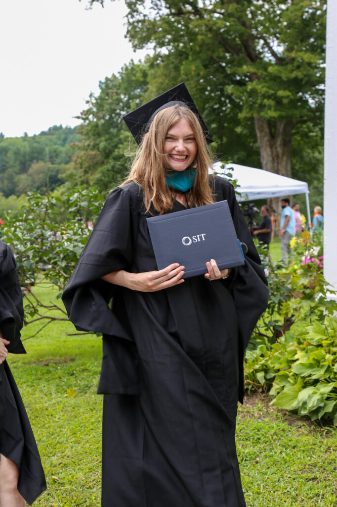 A young woman with long blond hair, wearing a black cap and gown, smiles as she displays an SIT diploma cover