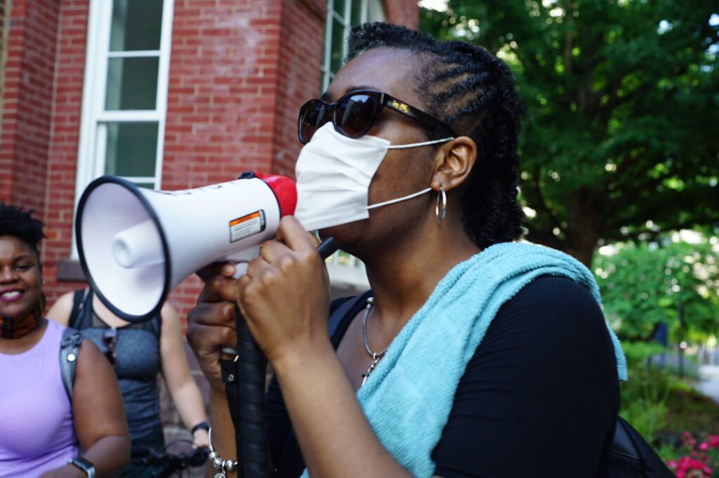 A Black woman wearing sunglasses and a surgical mask speaks into a megaphone.