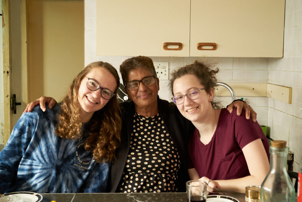 Three people smile toward the camera; the older woman in the middle has her arms around the shoulders of the two younger woman.