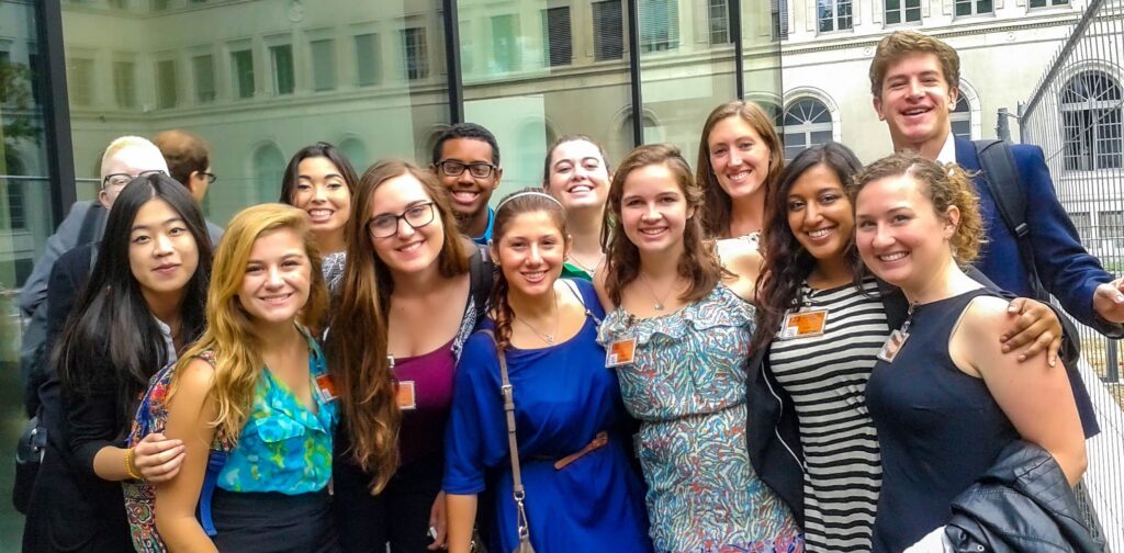 A diverse group of young people wearing business attire smile toward the camera. The background is a city setting.