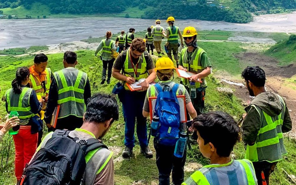 Students wearing green safety vests and yellow hardhats spread out along a lush green landscope. Some wear backpacks and other carry clipboards. One person in the middle of the group is writing something in a notebook.