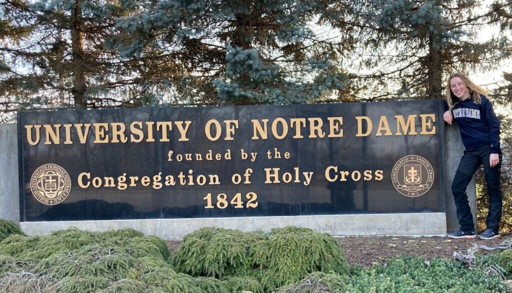 A person with shoulder-length blonder hair, wearing a blue track suits, stands next to a large University of Notre Dame.
