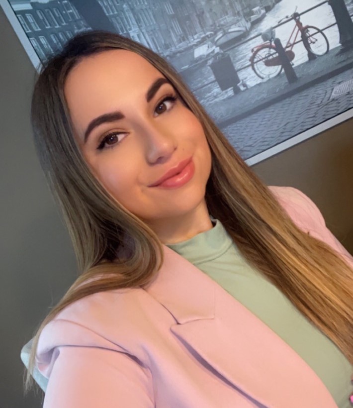 A young woman with long hair, wearing a pink jacket, smiles.