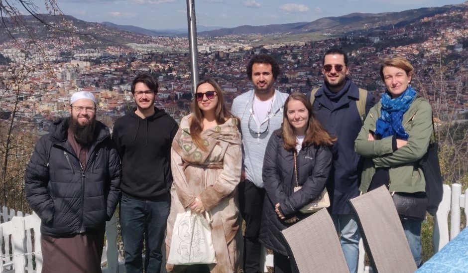 Seven people pose in a group against a backdrop of a city spread across low rolling hills.