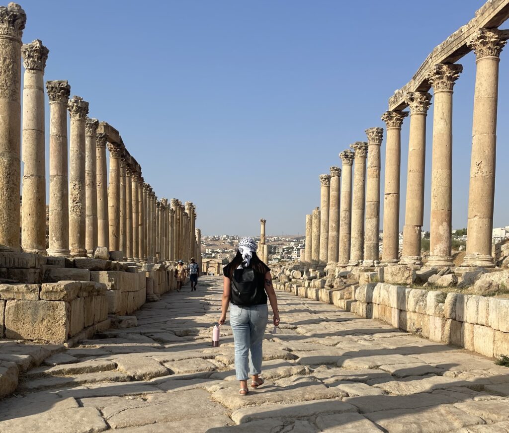 A person walks away from the camera, along a row of ancient columns along a street paved with ancient stones.