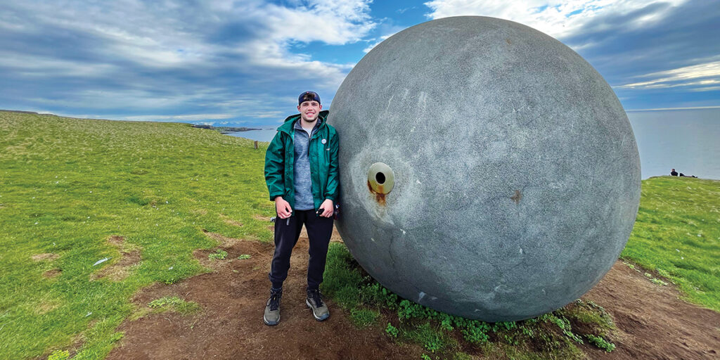 A person in green jack, black pants and a backward baseball cap stands next to a large round object.