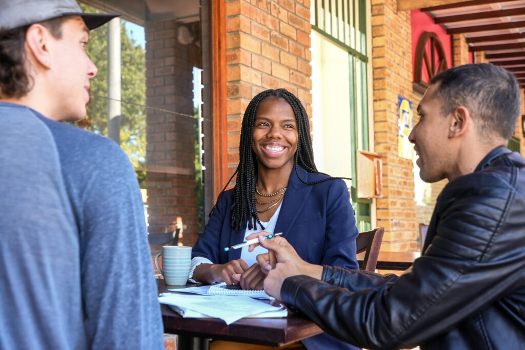 Three people have a conversation at an outdoor table. Their notebooks are spread out in front of them.