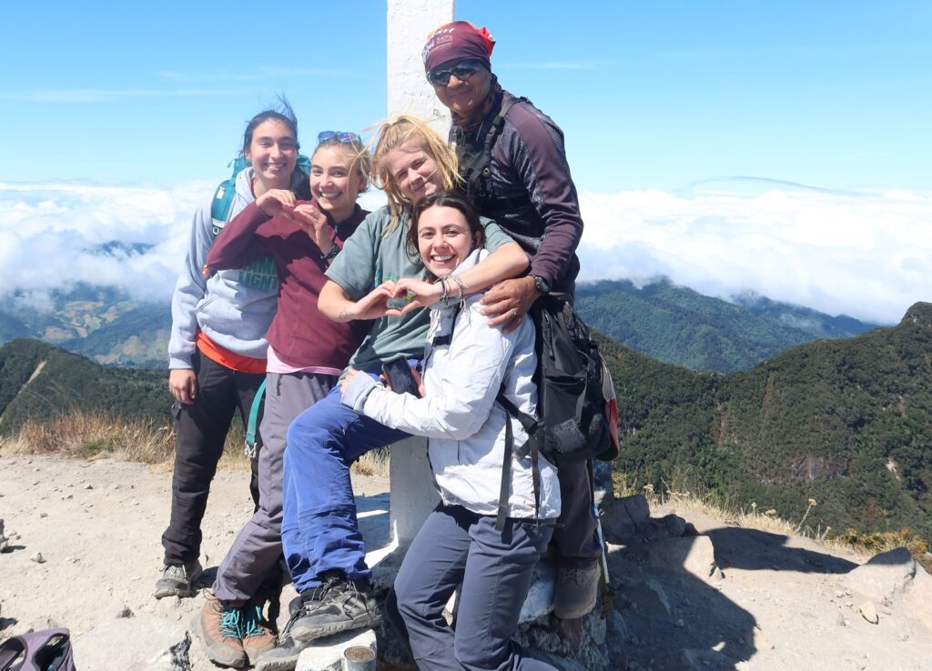 A group of five young people at the top of a mountain with mountains and clouds in the background