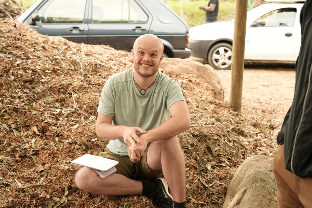 A young white man with a shaved head and a beard smiles. He is sitting atop what appears to be a pile of wood shavings. He has a notebook on his lap.