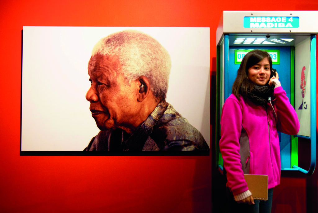 A student wearing a pink jacket listens to a recording in front of a large photo of Nelson Mandela.