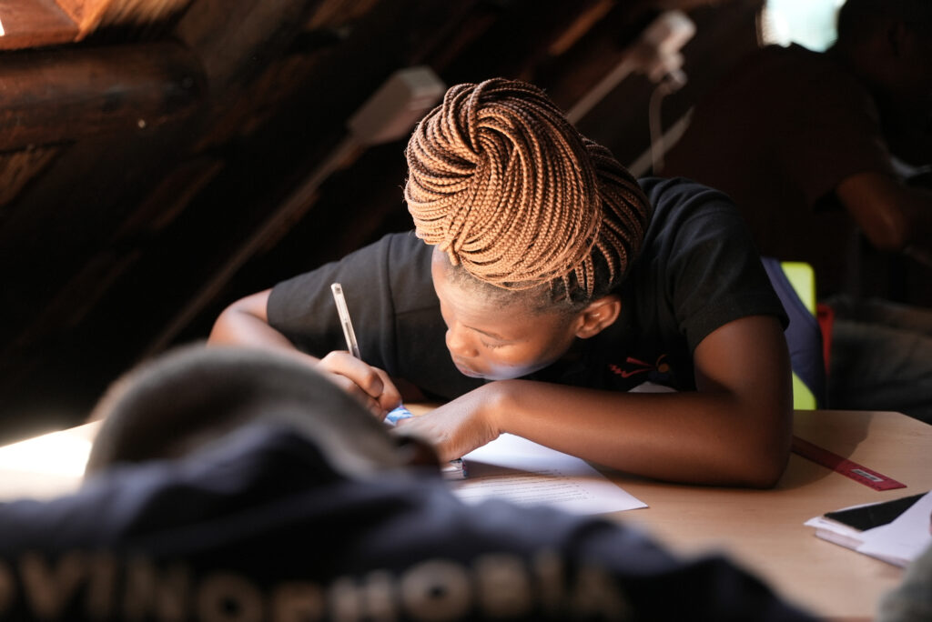 A student looks down as they write in a notebook. Their braided hair is arranged on top of their head.