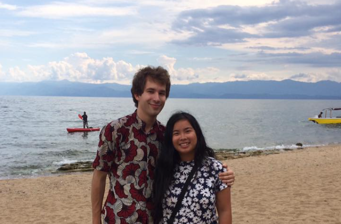 Two young people stand in front of a larger lake with mountains in the background. A board and a paddleboard are visible behind them.