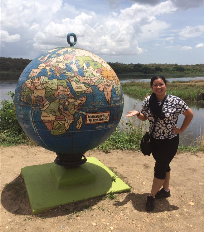 A person with long black hair, wearing a floral print shirt and black pants, stands next to a large globe.