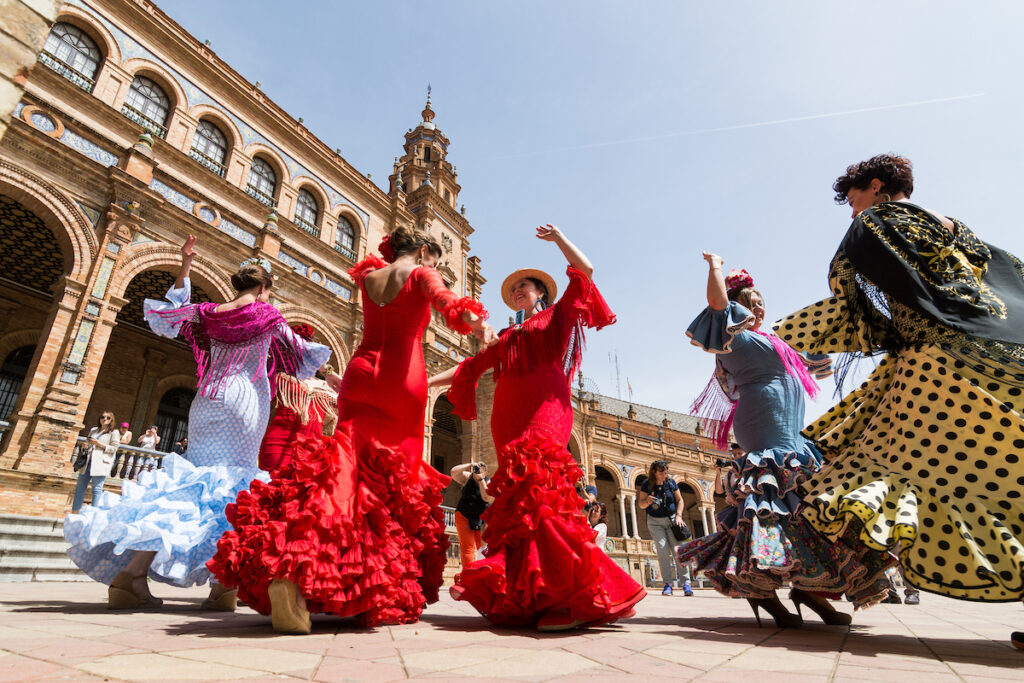Women in traditional dresses with dramatic ruffled skirts dance flamenco in a plaza in Seville.
