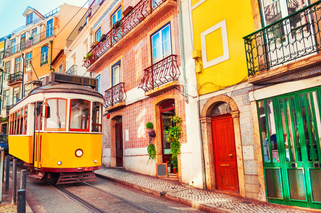 A vintage yellow tram car in front of colorful houses in Lisbon. Door and tile colors include bright red, green, yellow and orange.