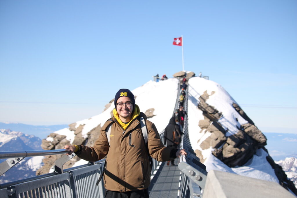 A young person smiles while standing on a narrow walkway leading up a hill topped by a Swiss flag. The person wears a University of Michigan cap and a brown parka.